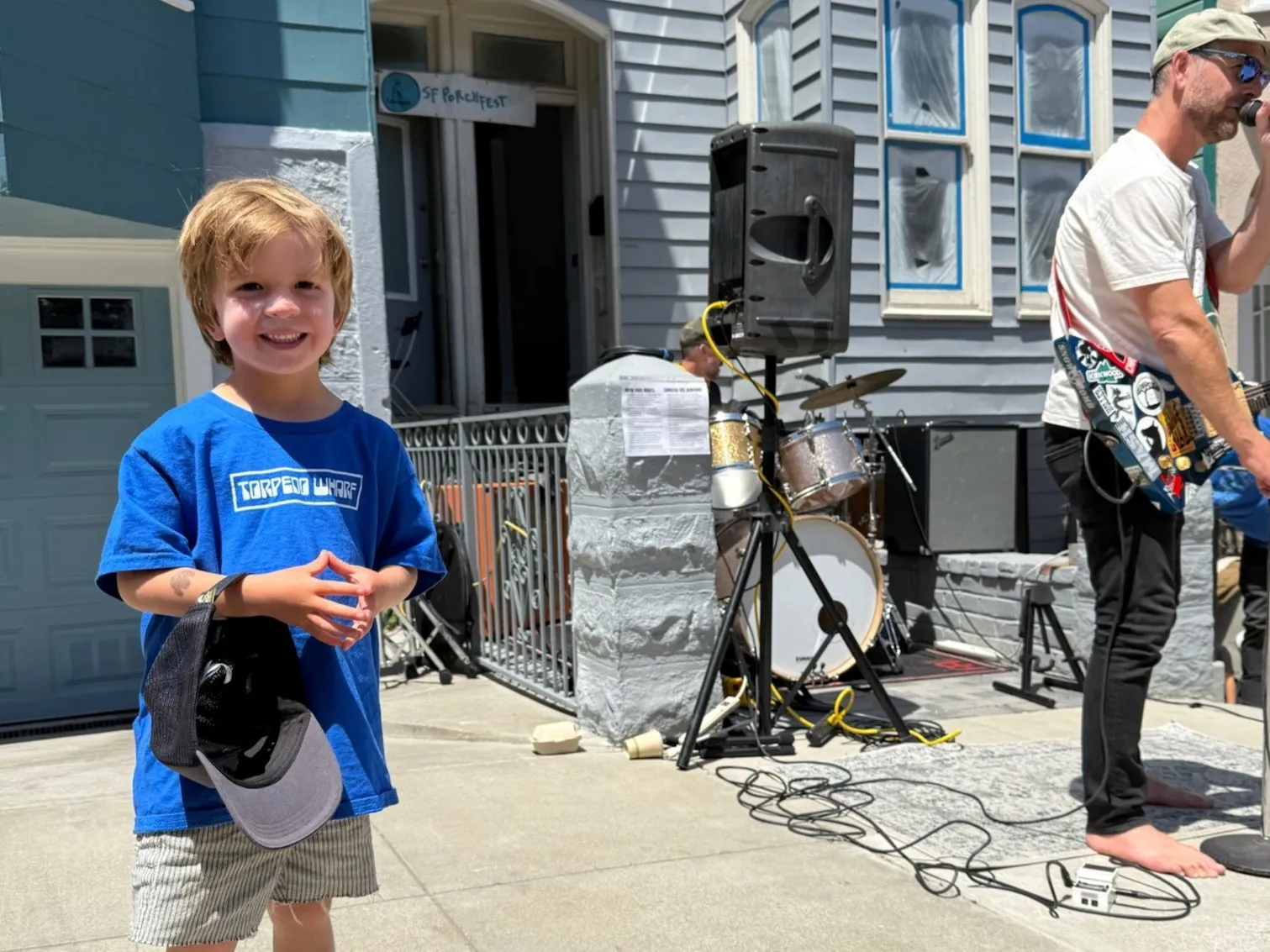 A young fan in a Torpedo Wharf shirt grinning while the band plays on the sidewalk at SF Porchfest